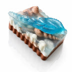 A decorative soap bar shaped like a slice of cake, featuring a brown base, white frosting-like layers, and translucent blue and clear sections with a seashell motif, shown on a white background.